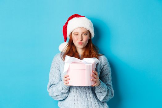 Winter holidays and Christmas Eve concept. Sassy redhead girl in sweater and Santa hat, holding New Year gift and looking at camera, standing against blue background.