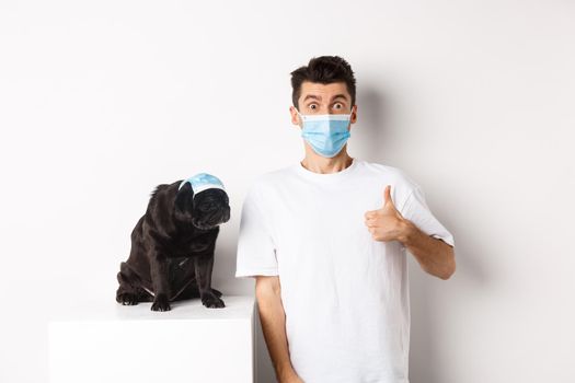 Covid-19, animals and quarantine concept. Handsome young man and small dog wearing face masks, owner showing thumb up in approval, praise super cool product, white background.