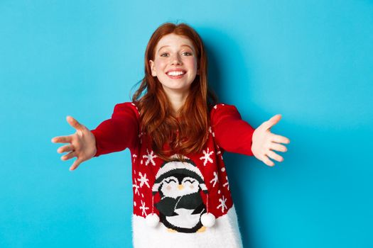 Winter holidays and Christmas Eve concept. Beautiful redhead girl in xmas sweater reaching for hug, extend hands for cuddles and smiling, standing in sweater against blue background.