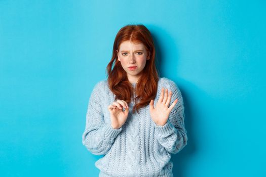 Reluctant redhead girl asking to stay away, shaking hand in rejection gesture, decline offer and grimacing displeased, standing over blue background.
