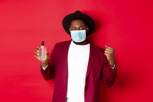 Covid-19, quarantine and holidays concept. Cheerful black man rejoicing of winning, achieve goal, showing antiseptic, wearing medical mask, standing against red background.