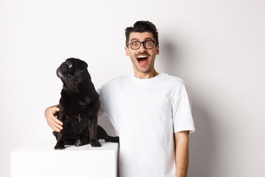 Image of amazed man staring at camera while his small dog looking left at copy space, standing over white background.