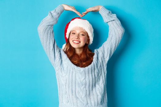 Winter holidays and Christmas Eve concept. Cute redhead teen girl in santa hat and sweater, making heart sign and smiling, wishing merry xmas, standing over blue background.