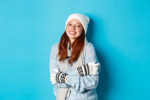 Winter and holidays concept. Smiling redhead girl in beanie, gloves and sweater getting warm after going outside, standing over blue background.