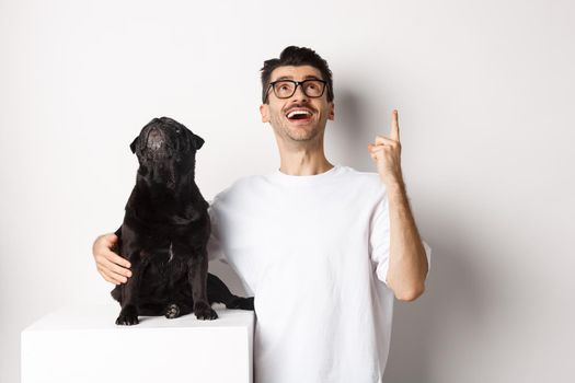 Image of happy young man and dog both looking up at promo, owner pointing finger at top, pug staring at loog, standing over white background.