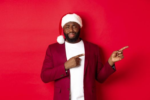 Christmas, party and holidays concept. Skeptical and unamused Black man looking with disdain, pointing fingers right at logo, standing in santa hat against red background.