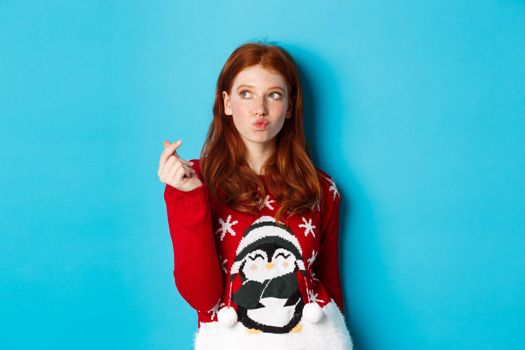 Winter holidays and Christmas Eve concept. Lovely redhead woman in xmas sweater, showing heart sign and thinking, looking upper left corner at logo, blue background.