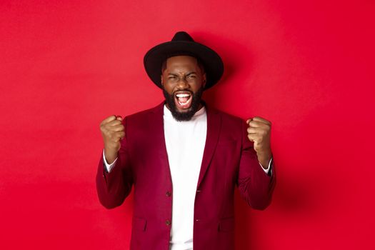 Satisfied lucky Black man celebrating victory, screaming from rejoice, winning prize, saying yes and fist pump, red background. Copy space