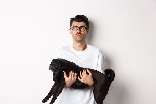 Confused hipster guy holding black cute pug and looking left, feeling puzzled or clueless. Dog owner pondering, carry dog in arms, white background.