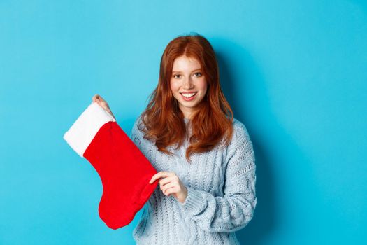 Winter holidays and gifts concept. Cute redhead girl in sweater showing Christmas stocking and smiling, celebrating New Year, standing over blue background.