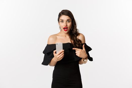 Online shopping concept. Stylish woman in black dress, wearing makeup, pointing finger at mobile phone with surprised emotion, standing over white background.