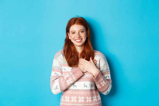 Image of beautiful redhead female model holding hands on heart and smiling, saying thank you, being grateful, standing over blue background.