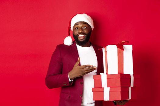 Christmas, New Year and shopping concept. Cheerful Black man secret santa holding xmas presents and smiling excited, bring gifts, standing against red background.