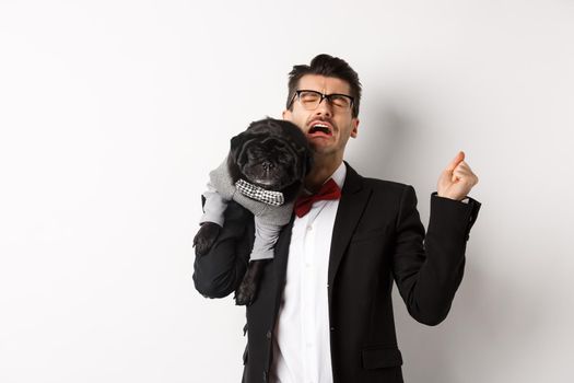 Sad man in suit, holding cute small dog on shoulder and crying with disappointed face, standing distressed over white background.