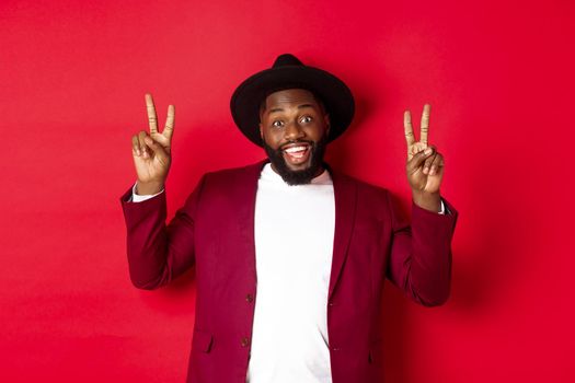 Fashion and party concept. Handsome Black man having fun, showing peace signs and smiling, standing in hat against red background.