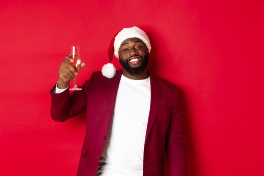 Christmas, party and holidays concept. Cheerful Black man saying cheers, raising glass of champagne and wishing happy New Year, standing against red background.