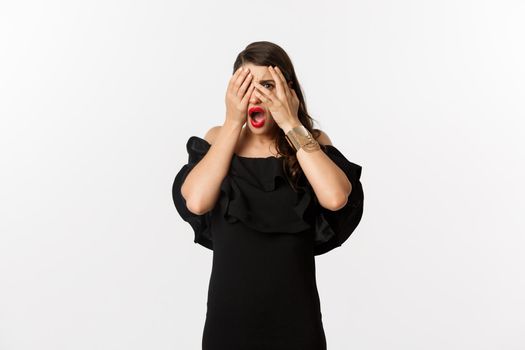 Fashion and beauty. Shocked young woman in black dress covering eyes, peeking through fingers at something embarrassing, cringe, standing over white background.