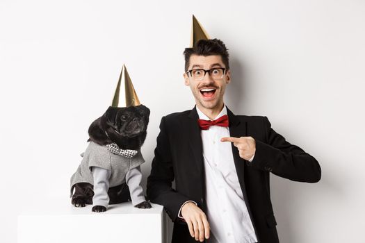 Cheerful man and cute black pug wearing party cones and suits, dog owner celebrating pet birthday, standing over white background.