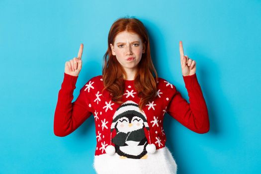 Winter holidays and celebration concept. Perplexed redhead girl in xmas sweater, frowning doubtful and pointing fingers up, showing advertisement, blue background.