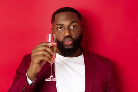 Close-up of funny Black man tasting champagne from glass, looking silly and pucker lips, celebrating new year, standing against red background.