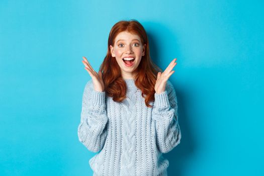Surprised and happy redhead girl clap hands and staring at camera, smiling amazed, standing against blue background.