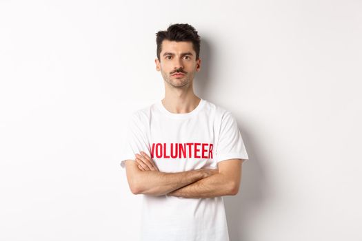 Serious young male volunteer in white t-shirt, holding arms crossed on chest, looking at camera, ready to help.
