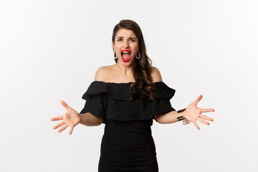 Fashion and beauty. Angry woman in black dress, shouting mad and shaking hands, grimacing outraged at camera, standing over white background.