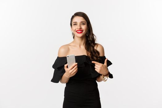 Online shopping concept. Stylish woman in black dress, wearing makeup, pointing finger at mobile phone with satisfied smile, standing over white background.