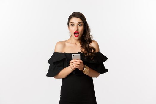 Happy young glamour woman in black dress, holding mobile phone and looking surprised, standing over white background.