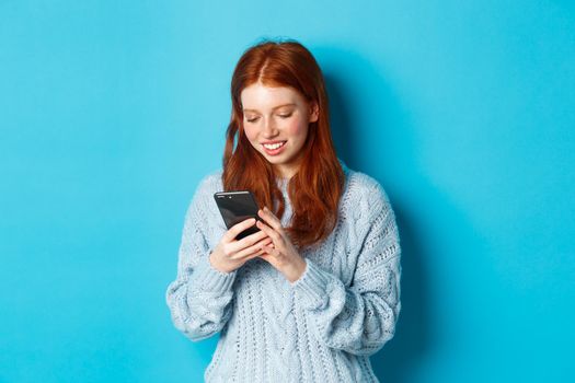 Young teenage redhead girl reading message on smartphone and smiling, using mobile phone and standing over blue background.