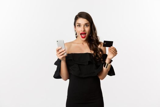 Online shopping concept. Fashionable woman in black dress, holding credit card with smartphone, looking excited, standing over white background.