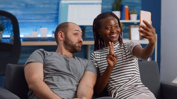 Cheerful interracial couple taking pictures with smartphone sitting together on living room couch. Multi ethnic married people having fun with selfies and modern device at home