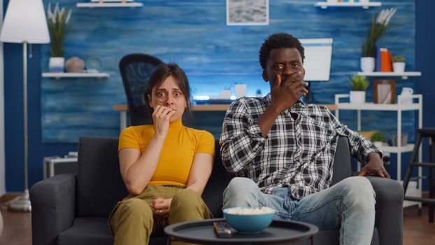 POV of shocked interracial couple watching scary movie on television at home. Multi ethnic husband and wife being afraid and covering eyes while looking at camera in living room