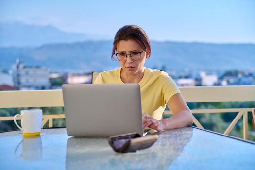 Serious middle aged woman with laptop at table on outdoor balcony. Concentrated 40s mature business woman working remotely. Freelance, technology, work from home office, online training, blog, vlog