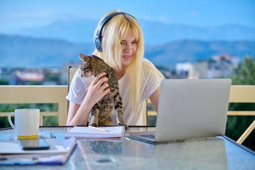Female student studies at home online using laptop. Teenager sitting on outdoor balcony with pet cat in headphones, school notebooks, looking at screen. E-learning, modern technologies in education