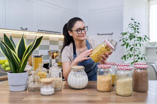 Food, grocery storage, smiling woman looking at the camera in the kitchen, with a jar of pasta in hand. Organization of storage of bulk products in glass jars and containers, pantry