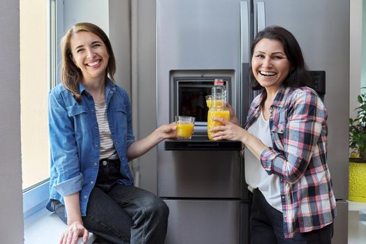 Two middle-aged women drinking orange juice. Smiling female friends talking in the kitchen near the refrigerator. People, food, drinks, diet, rest, communication, lifestyle concept