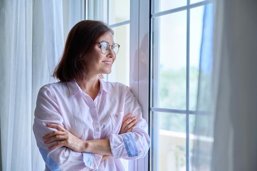 Portrait of a mature confident woman looking out the window. Middle-aged female wearing glasses with crossed arms, calm, smiling, resting