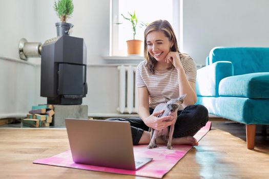 Middle-aged smiling woman with laptop and cat on the floor. Positive female sitting on a yoga mat in a lotus position with a pet in her arms looking at a laptop screen using a video call