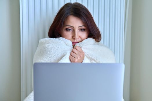 A frozen mature woman working at home with a laptop, warming herself with a blanket and heating radiator. Autumn winter cold season