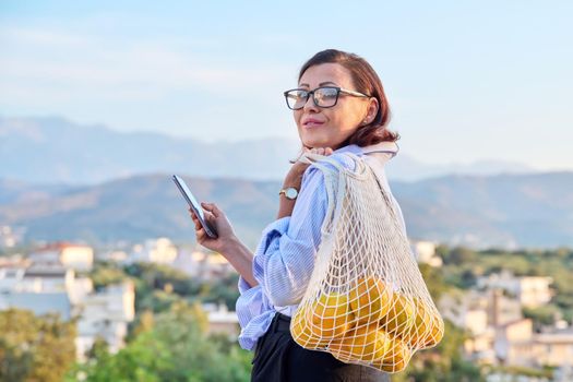 Portrait of beautiful middle-aged woman with smartphone trending grid of oranges outdoors. Female with phone and eco mesh shopping bag with organic oranges, mountains village sunset background, trends