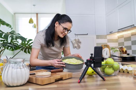 Technologies in everyday life, 40s woman preparing apple pie at home in kitchen, with smartphone on tripod using video call for communication. Lifestyle, eating at home, technology, people concept