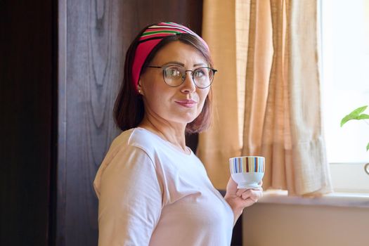 Portrait of middle aged woman with glasses headband at home in kitchen with cup of coffee, copy space. Female housewife looking at camera, lifestyle, rest, morning coffee, mature people