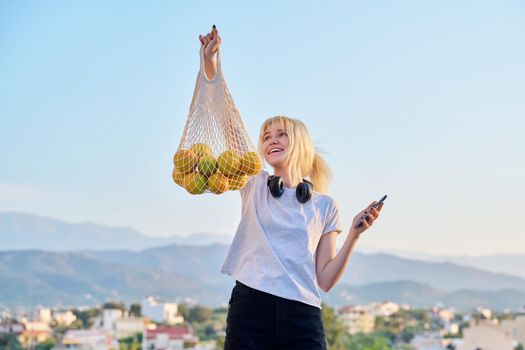 Happy female teenager with eco-friendly mesh bag of farm organic oranges, sunset village mountains. Food trends, buying natural farm fruits, using reusable non-plastic shoppers, fashion and ecology