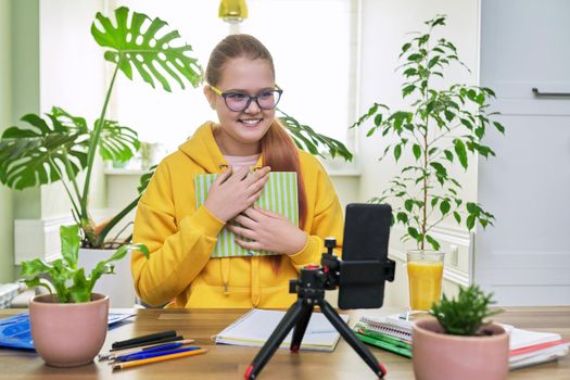 Teenage girl looking at webcam using smartphone for an online lesson, reading textbook book. Student 12, 13 years old, studying remotely at home, e-learning, virtual lessons, distance learning