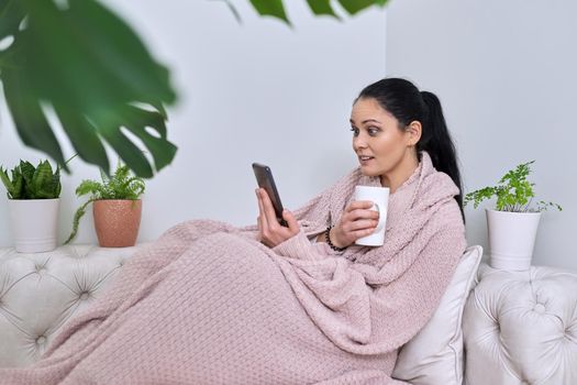 Middle-aged woman having a rest at home in winter fall season, surprised female looking at phone screen. Under warm knitted blanket with smartphone and cup of hot tea