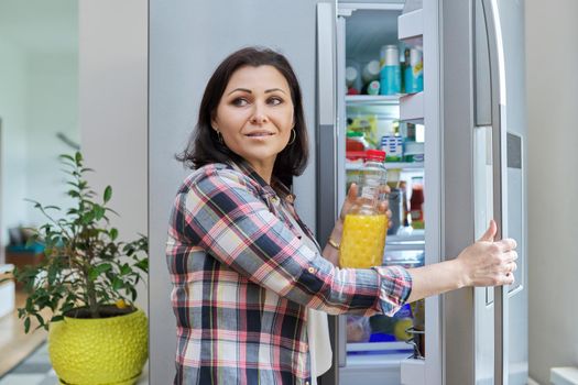 Woman opens refrigerator at home in kitchen, holding bottle of orange juice. People, food, drinks, lifestyle, home, kitchen, refrigerator, groceries concept