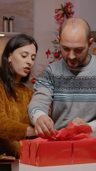 Couple preparing gift box for family on christmas eve festivity. Festive man and woman wrapping paper and ribbon on present for seasonal celebration and winter holiday. People decorating