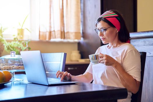 Mature woman relaxing at home in living room with laptop and cup of coffee. 40s female enjoying drink, rest, reading and watching video on laptop. Technology, lifestyle, home life, mature people