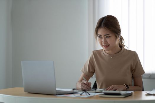 portrait of smiling woman making notes while taking part in webinar at tabletop with laptop in office.